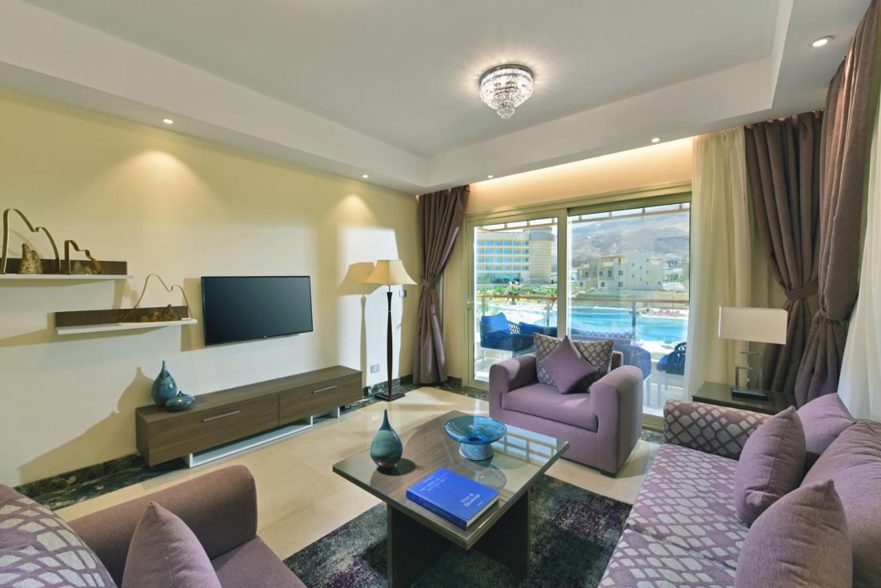 Tolip Resort El Galala Majestic Ain Sukhna Buitenkant foto The photo shows a modern living room that features a stylish and cozy design. There are two purple sofas with decorative cushions arranged around a coffee table. The coffee table has a blue book and a decorative item on it. A flat-screen TV is mounte