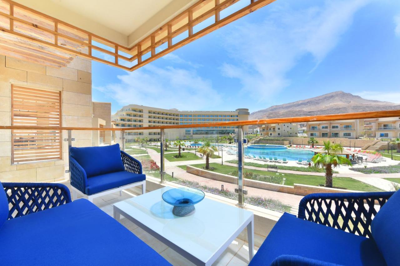 Tolip Resort El Galala Majestic Ain Sukhna Buitenkant foto The photo showcases a balcony view featuring two blue chairs and a table with a decorative bowl. The balcony overlooks a landscape that includes a swimming pool and manicured lawns. In the background, there are multiple buildings and palm trees, all 