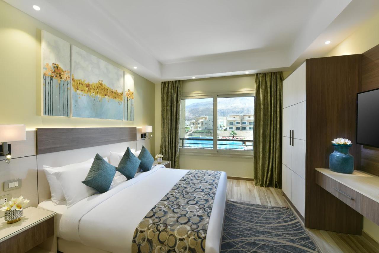 Tolip Resort El Galala Majestic Ain Sukhna Buitenkant foto The photo shows a modern hotel room. It features a large bed with white bedding and several decorative pillows in shades of teal. Above the bed, there are two pieces of wall art with a landscape theme. The room has a large window with green curtains,