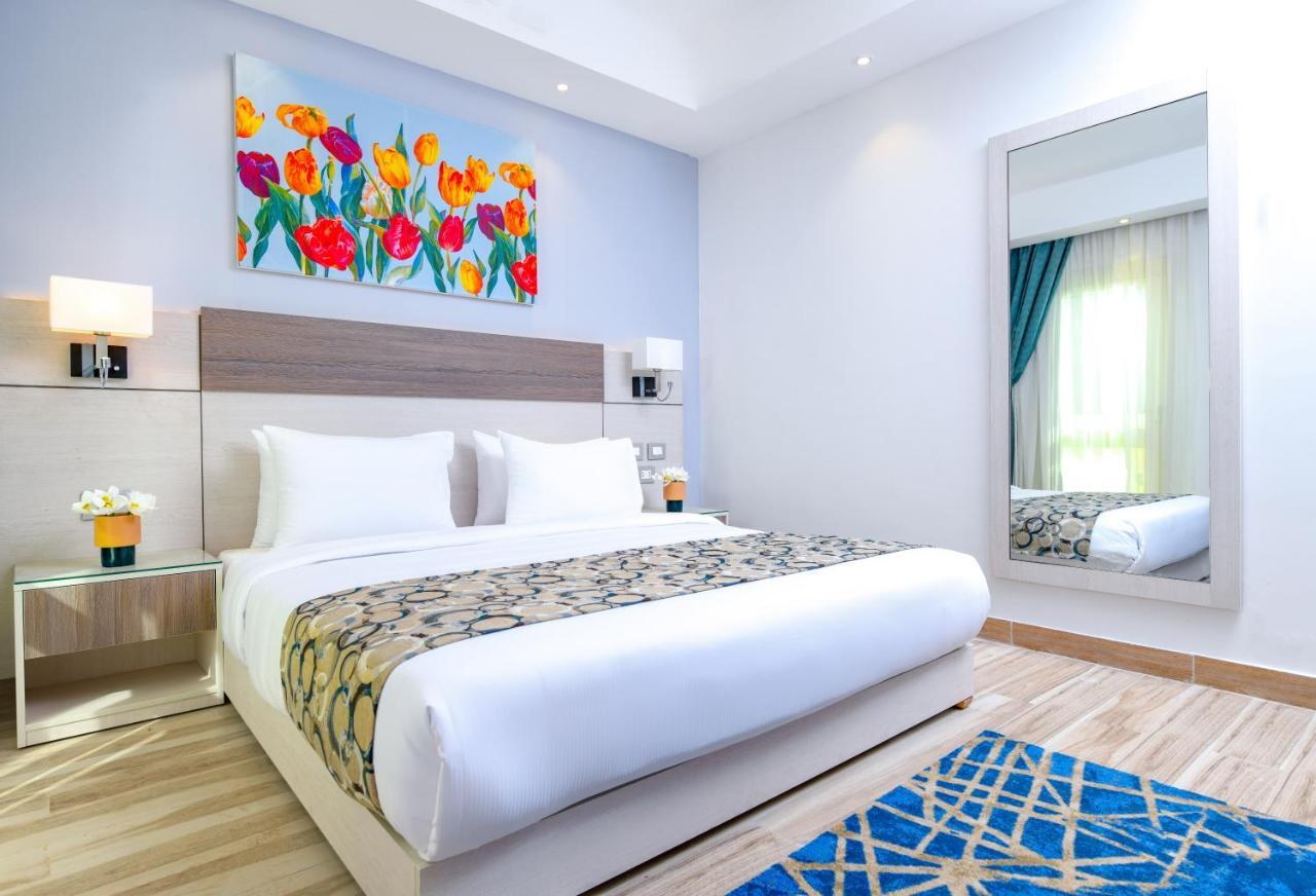 Tolip Resort El Galala Majestic Ain Sukhna Buitenkant foto The photo shows a modern hotel bedroom. It features a large bed with white linens and a decorative throw blanket. The room has light wooden flooring and is bright, with ample natural light coming from a window. There is a colorful painting of flowers