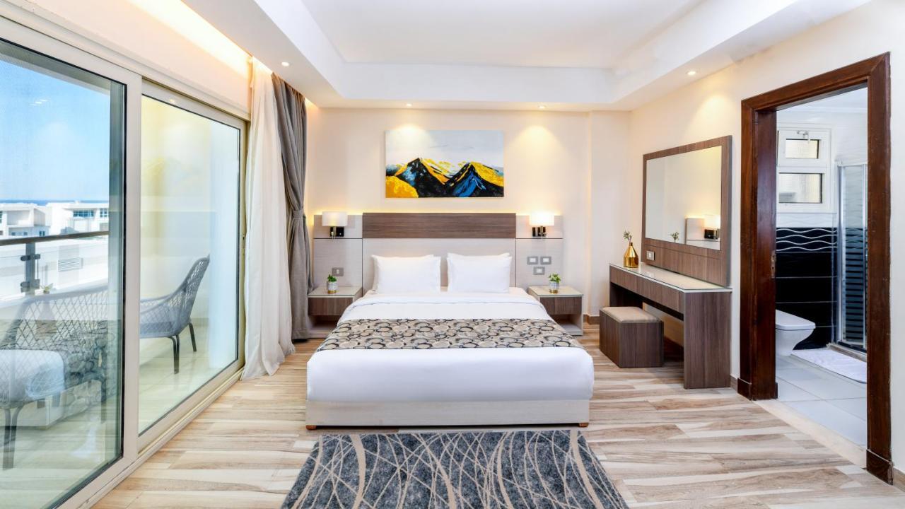 Tolip Resort El Galala Majestic Ain Sukhna Buitenkant foto The photo shows a modern hotel room featuring a large bed with white linens and a decorative throw. The room has a neutral color palette, accented with warm lighting. There is a piece of artwork above the bed depicting mountains. To the left, there a