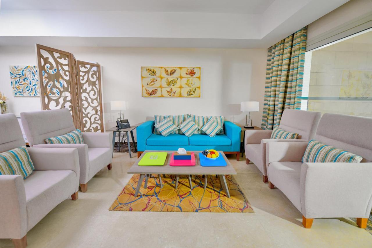 Tolip Resort El Galala Majestic Ain Sukhna Buitenkant foto The photo shows a stylish and colorful living room. It features a bright blue sofa with patterned cushions, complemented by two light gray armchairs. In front of the sofa, there's a wooden coffee table with vibrant trays in pink, green, and yellow. A
