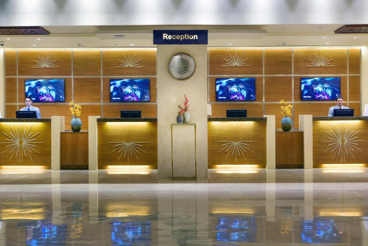 Tolip Resort El Galala Majestic Ain Sukhna Buitenkant foto The image shows a hotel reception area. There are several reception desks lined up against a wall with decorative paneling. Each desk has a staff member, and above the desks, there are screens displaying information. A clock is visible on the wall, a