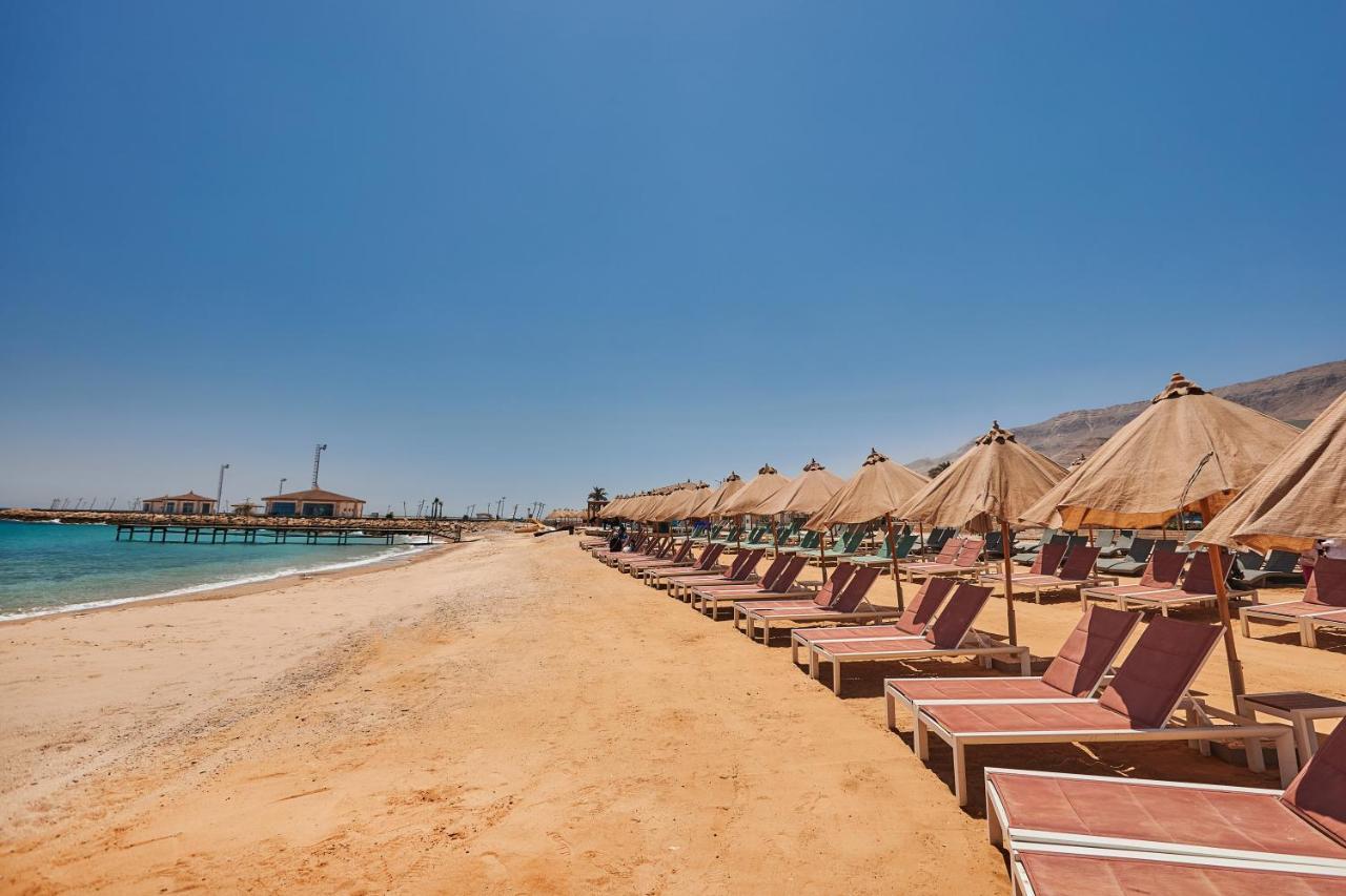 Tolip Resort El Galala Majestic Ain Sukhna Buitenkant foto The photo shows a scenic beach with a stretch of golden sand under a clear blue sky. There are several beach loungers arranged in rows, each accompanied by thatched umbrellas providing shade. In the background, there is a wooden pier extending into t