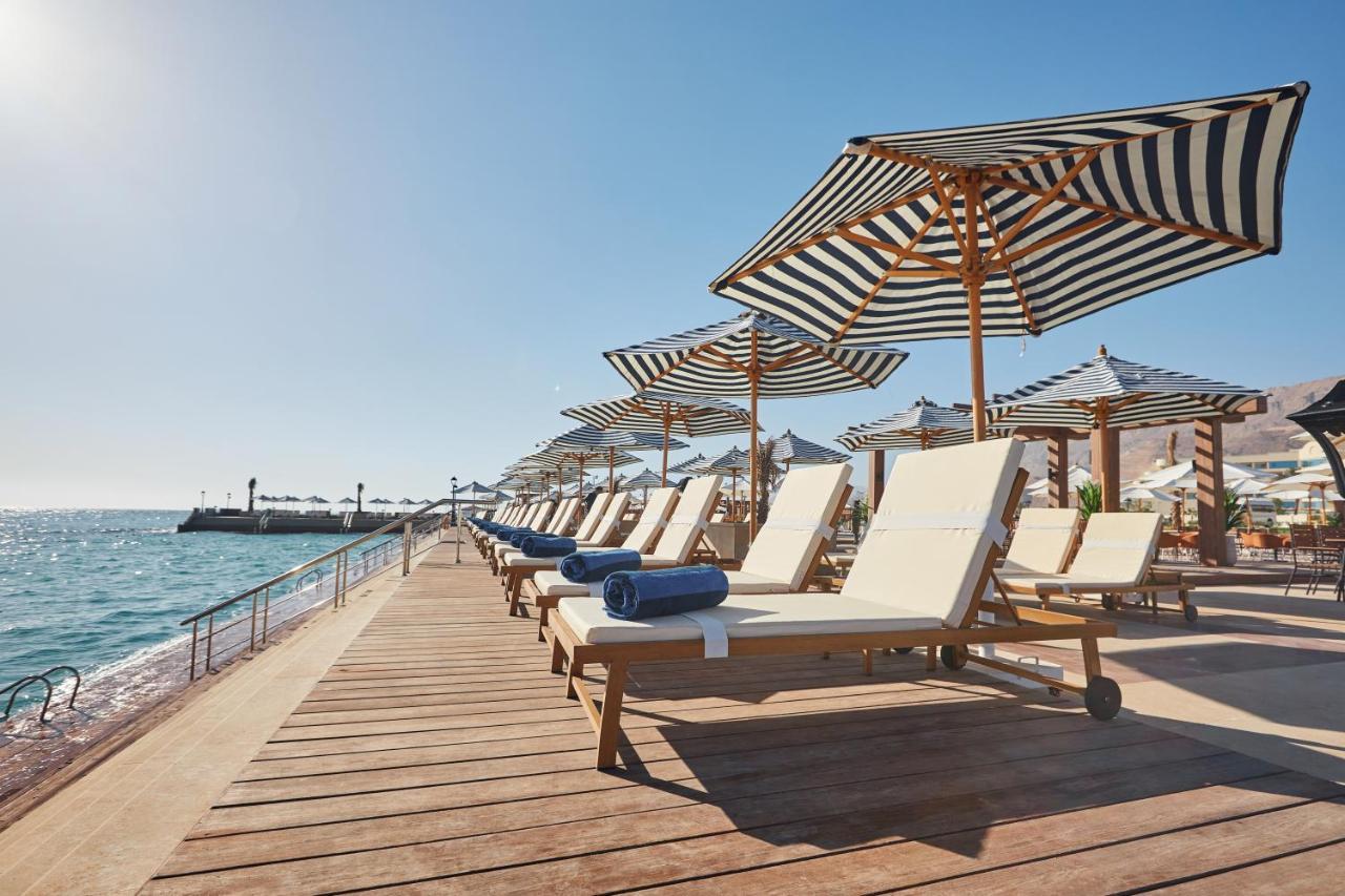 Tolip Resort El Galala Majestic Ain Sukhna Buitenkant foto The photo depicts a serene beachside setting featuring a row of sun loungers arranged on a wooden deck. Each lounger is equipped with a folded towel and shaded by striped umbrellas. In the background, the water sparkles under bright sunlight, and the