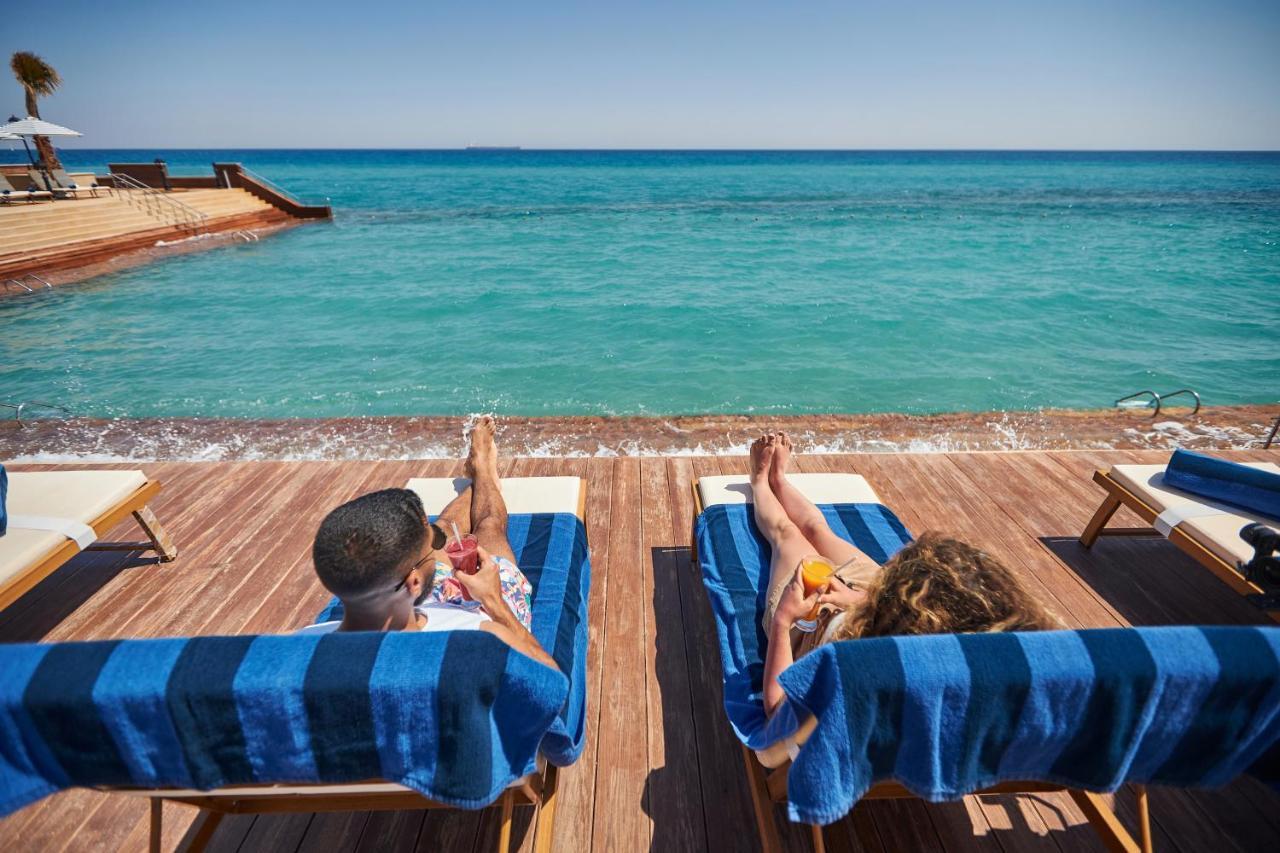 Tolip Resort El Galala Majestic Ain Sukhna Buitenkant foto The photo depicts two people relaxing on lounge chairs by the waterfront. They are positioned on a wooden deck with a view of the clear, turquoise water. Both individuals are reclining, enjoying the sun, and each has a drink in hand. The scene convey