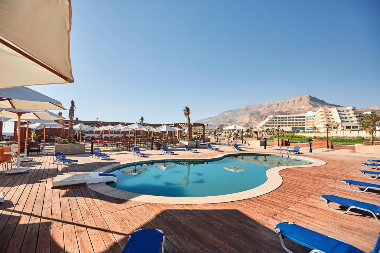 Tolip Resort El Galala Majestic Ain Sukhna Buitenkant foto The photo shows a swimming pool area surrounded by sun loungers and umbrellas. The pool has a unique, curvy shape and is situated on a wooden deck. In the background, there are several buildings, likely part of a resort or hotel, and a mountainous la