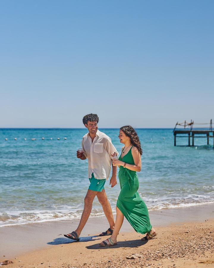 Tolip Resort El Galala Majestic Ain Sukhna Buitenkant foto The photo shows a couple walking along a beach. The man is wearing a white short-sleeved shirt and green shorts, while the woman is dressed in a flowing green dress. They both appear to be enjoying the pleasant weather, with the ocean in the backgrou