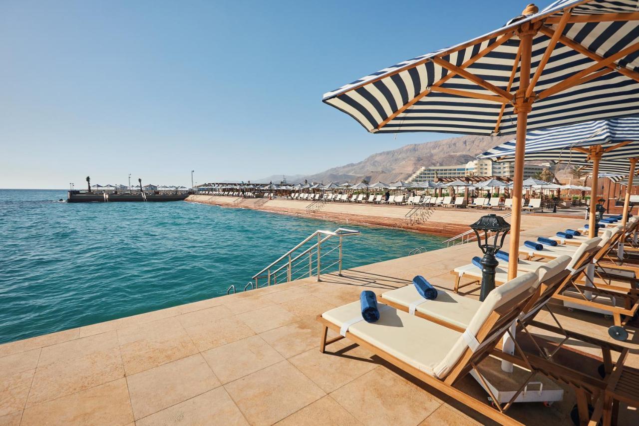 Tolip Resort El Galala Majestic Ain Sukhna Buitenkant foto The photo shows a scenic beachside area with clear blue water. In the foreground, there are several sun loungers set up on a tiled surface, with blue towels neatly rolled on each one. The loungers are shaded by striped umbrellas. In the distance, a w