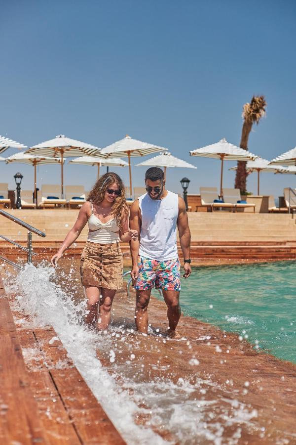 Tolip Resort El Galala Majestic Ain Sukhna Buitenkant foto The photo depicts a couple walking along a poolside area. They are enjoying a sunny day near a swimming pool, with splashes of water around them. The woman is wearing a light-colored top and a patterned skirt, while the man is dressed in a sleeveless