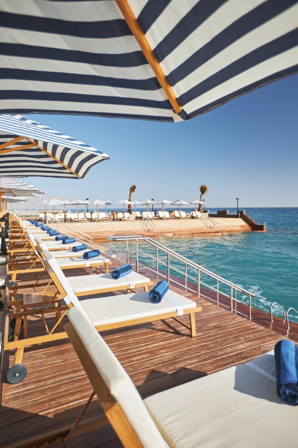 Tolip Resort El Galala Majestic Ain Sukhna Buitenkant foto The photo depicts a serene outdoor area by the waterfront. There are several lounge chairs arranged in a row, each equipped with blue towels. Large, striped umbrellas provide shade over some of the chairs. In the background, there is a wooden deck th