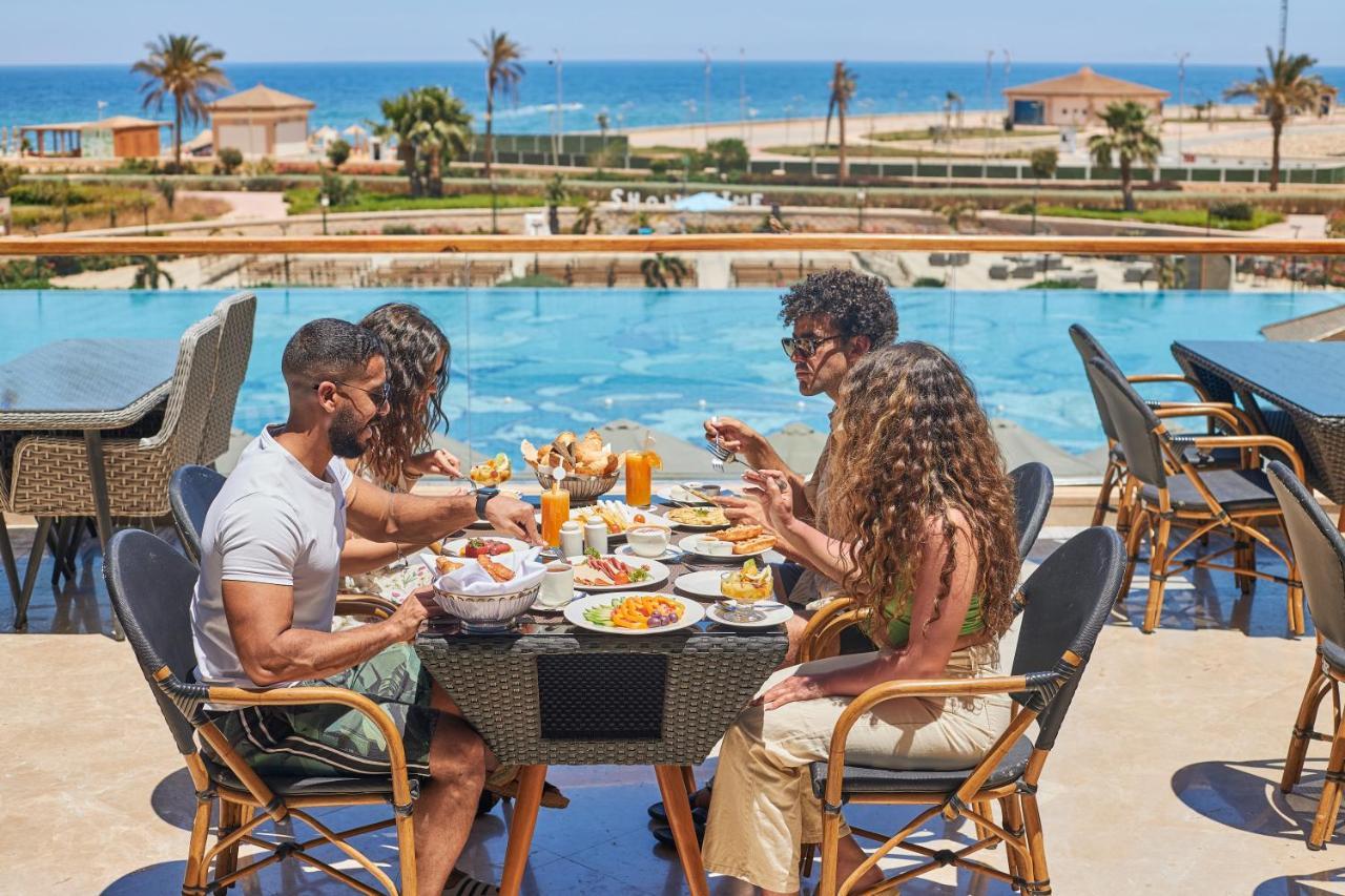 Tolip Resort El Galala Majestic Ain Sukhna Buitenkant foto The photo shows a group of five people enjoying a meal together outdoors, likely at a restaurant or cafe. They are seated at a table with a beautiful view of a swimming pool and the sea in the background. The table is filled with various dishes, and 