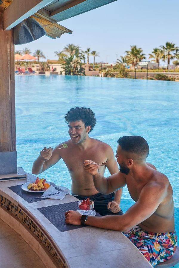 Tolip Resort El Galala Majestic Ain Sukhna Buitenkant foto The photo features two young men enjoying a meal together. They are sitting at a poolside table with a clear blue swimming pool in the background. The men are shirtless, suggesting a casual, relaxed atmosphere, likely in a warm environment. One man i