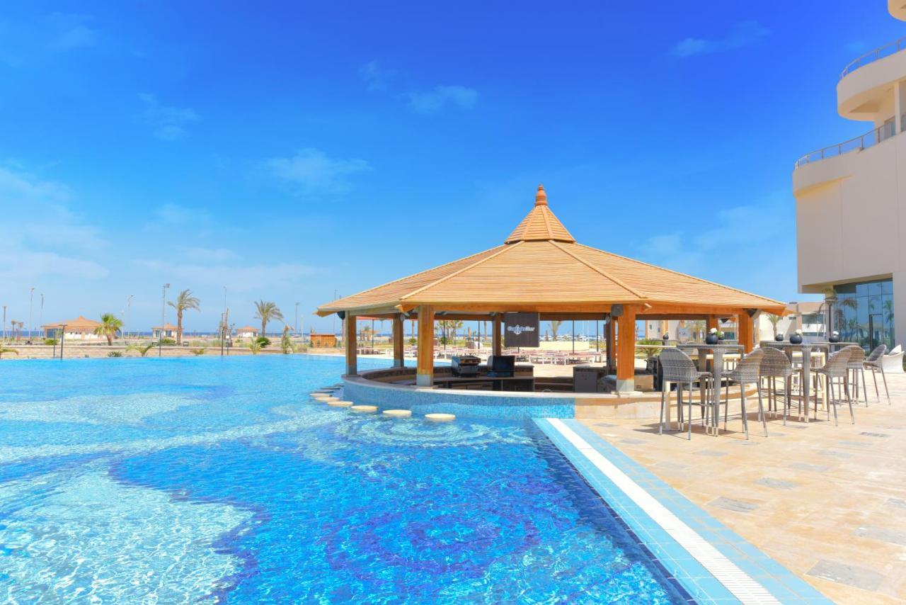 Tolip Resort El Galala Majestic Ain Sukhna Buitenkant foto The photo shows a beautiful outdoor setting featuring a large swimming pool with clear blue water. In the center, there is a thatched-roof gazebo, which serves as a shaded seating area. Surrounding the gazebo are lounge chairs and a bar area with hig