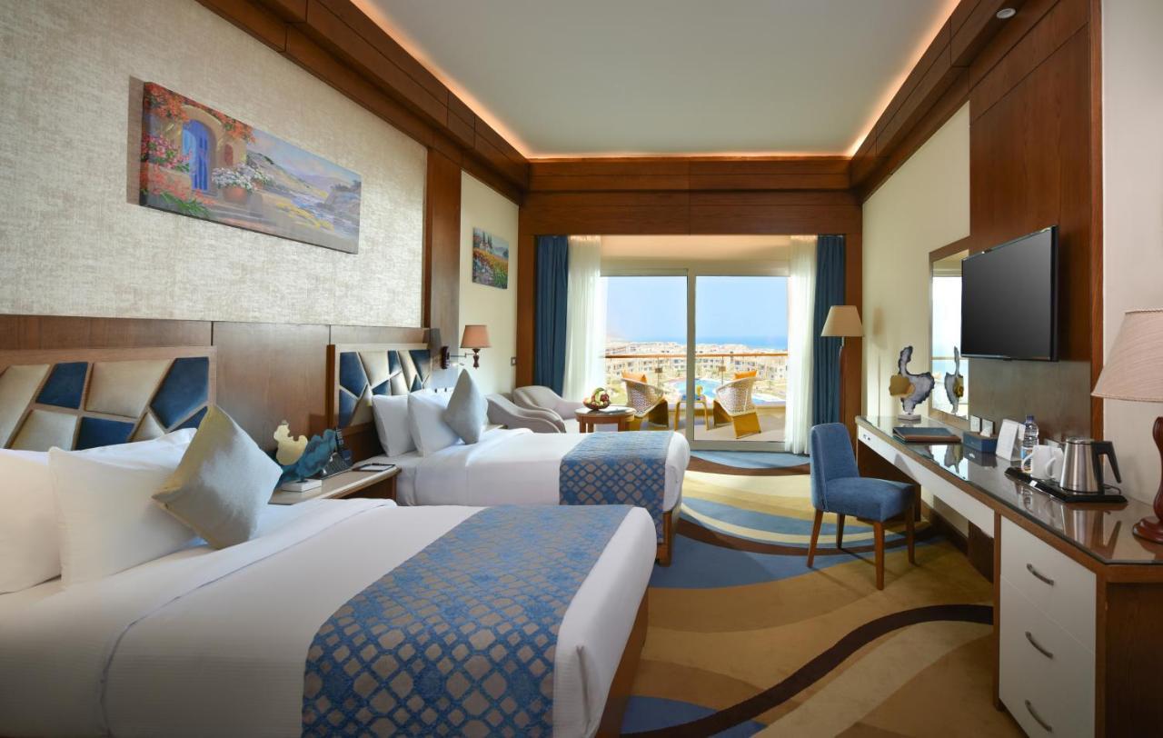 Tolip Resort El Galala Majestic Ain Sukhna Buitenkant foto The photo shows a well-decorated hotel room featuring two beds with decorative pillows. The room has wooden accents and modern furnishings, including a desk, a television, and lamps. Large windows flood the space with natural light, offering a view o