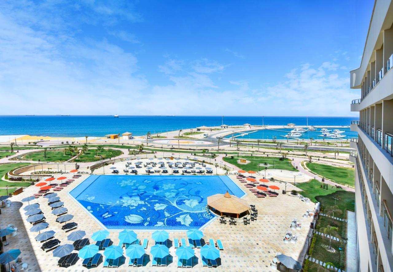 Tolip Resort El Galala Majestic Ain Sukhna Buitenkant foto The photo shows a beautiful outdoor view of a resort area featuring a large swimming pool with a decorative underwater design. Surrounding the pool are numerous sun loungers and umbrellas in a variety of colors. In the background, there is a scenic c