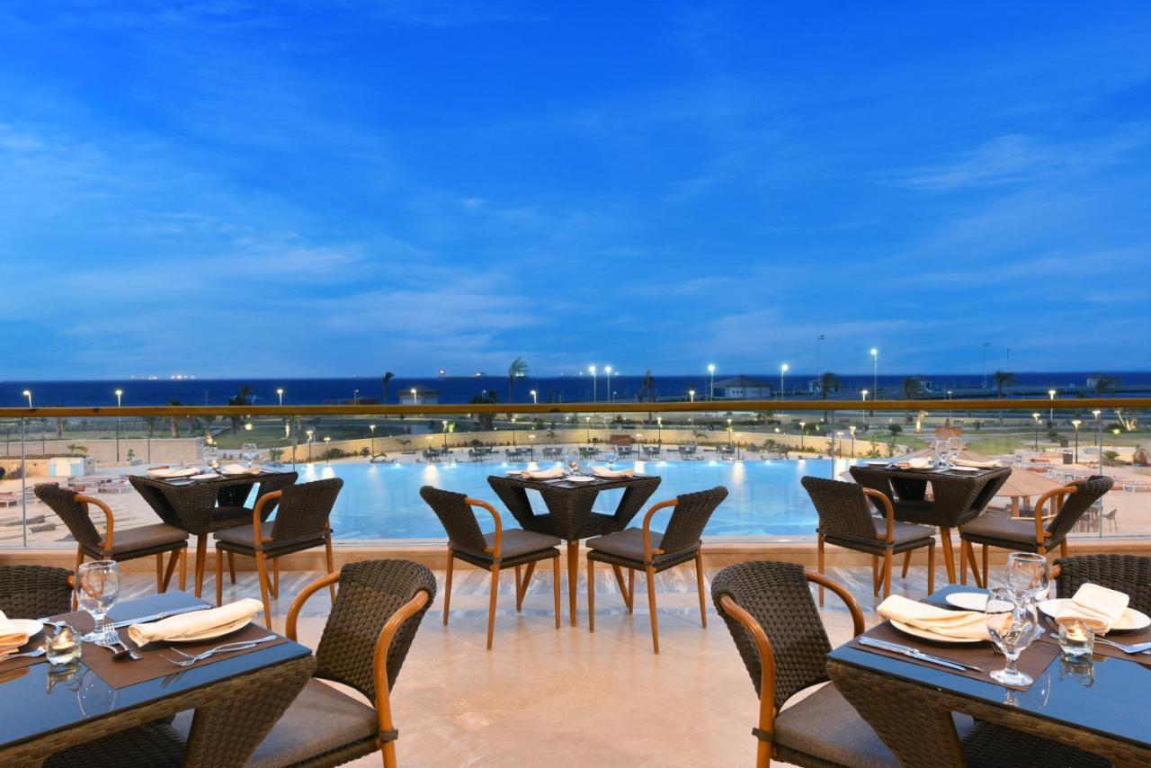 Tolip Resort El Galala Majestic Ain Sukhna Buitenkant foto The photo shows an outdoor dining area with several neatly arranged tables and chairs. The tables are elegantly set with cutlery and plates, suggesting a fine dining experience. In the background, there is a large swimming pool that reflects the blue