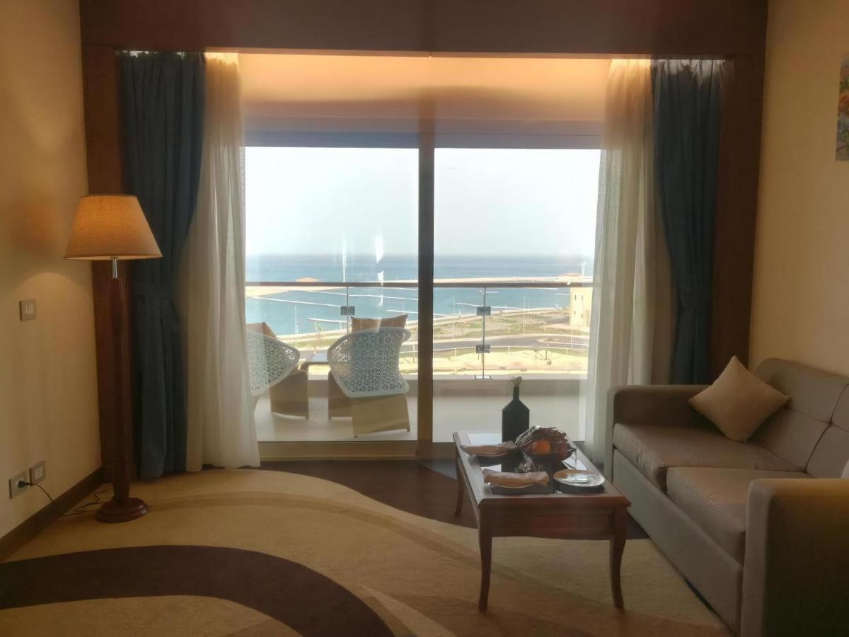 Tolip Resort El Galala Majestic Ain Sukhna Buitenkant foto The photo shows a stylish interior of a room with a large window overlooking the sea. The window features sheer and thick curtains and is framed by a balcony with two chairs visible outside. Inside, there is a comfortable-looking sofa, a coffee table