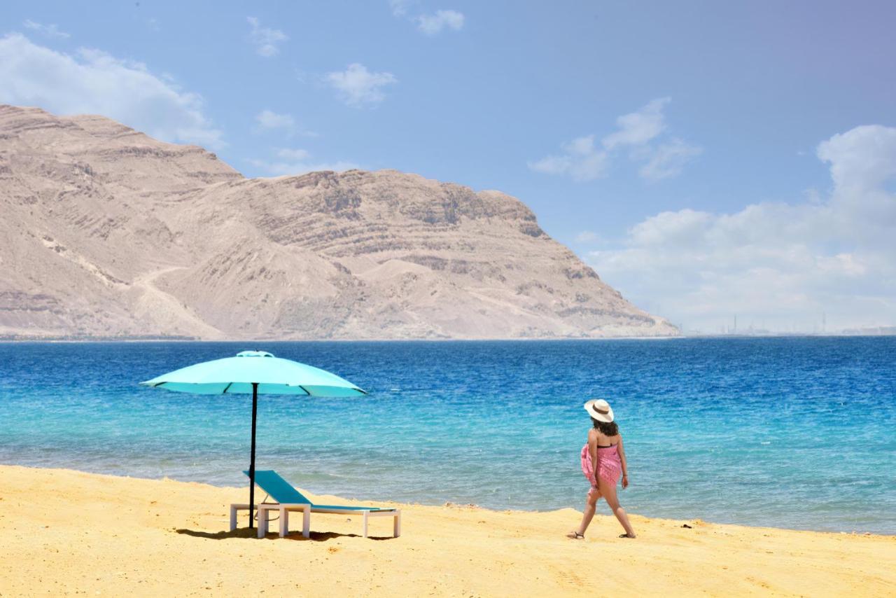 Tolip Resort El Galala Majestic Ain Sukhna Buitenkant foto The photo depicts a serene beach scene. In the foreground, there is a sandy shore with a single beach chair and an umbrella in a light blue color. A person dressed in a pink outfit is walking along the shoreline, enjoying the beach atmosphere. In the